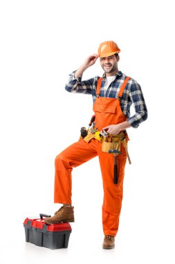 Smiling handyman in orange overall and hard hat leaning on tool box isolated on white