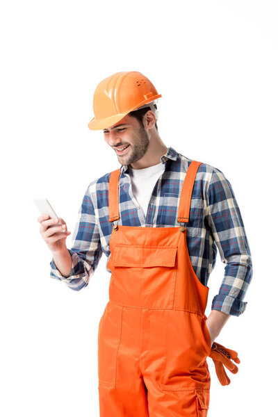 Smiling handyman in orange overall and helmet using smartphone isolated on white