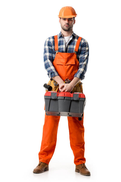Confident handyman in orange overall and hard hat holding tool box isolated on white