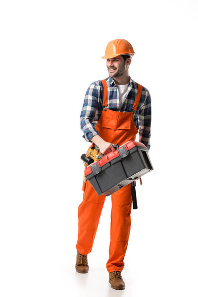 Smiling builder in orange overall and helmet carrying tool box isolated on white