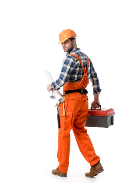 Young builder in orange overall and helmet carrying tool box and blueprint isolated on white