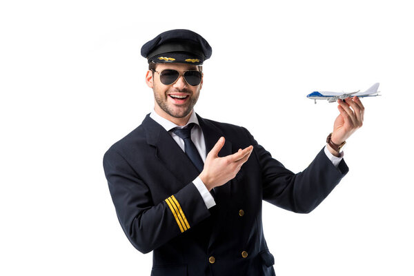 portrait of smiling bearded pilot in uniform pointing at toy plane in hand isolated on white
