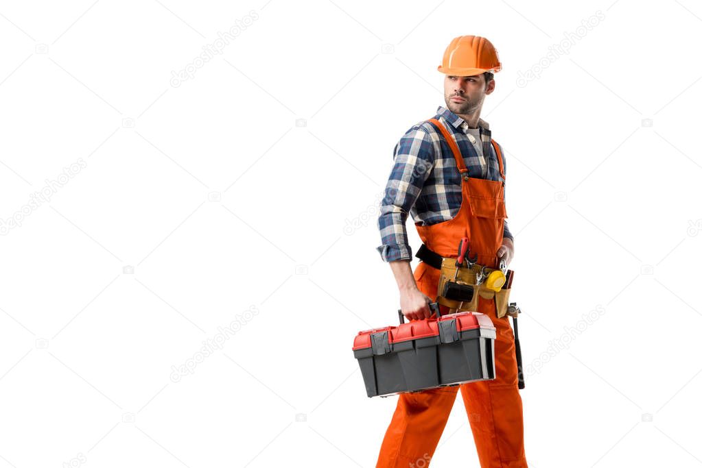 Young builder in orange overall and hard hat carrying tool box isolated on white