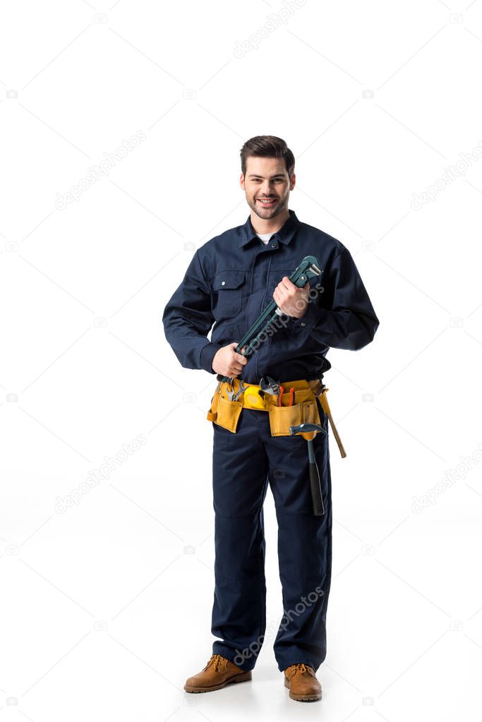 Confident handyman wearing uniform with tool belt and holding wrench isolated on white