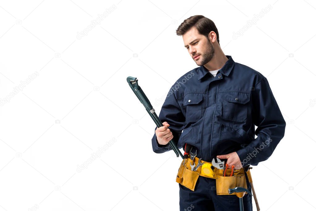 Bearded sanitary engineer wearing uniform with tool belt and looking at wrench isolated on white