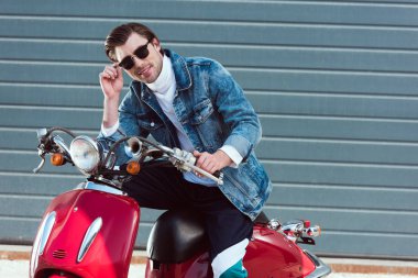 Kameraya bakarak vintage kırmızı scooter üzerinde gülümseyen genç adam