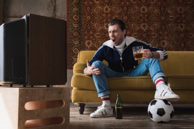 expressive young man in vintage clothes with ball and mug of beer watching soccer on old tv and shouting  clipart