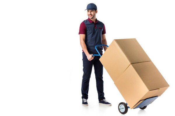smiling delivery man in uniform pushing hand truck with cardboard boxes isolated on white