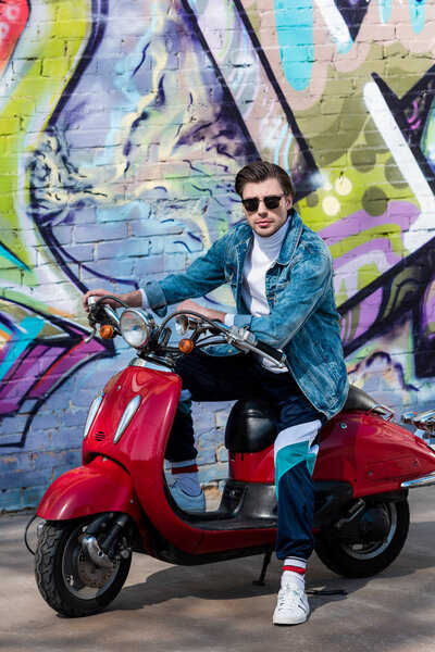 attractive young man on vintage red scooter in front of brick wall with graffiti