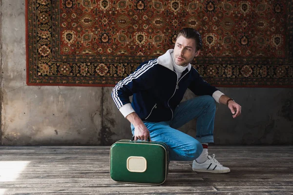 handsome young man with vintage little suitcase sitting squat in front of rug hanging on wall