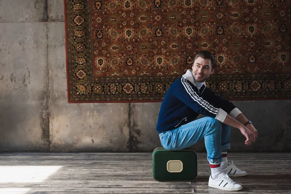 Happy Young Man Sitting Vintage Little Suitcase Front Rug Hanging — Stock Photo, Image