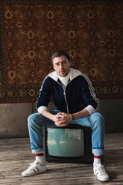 handsome young man in vintage clothes sitting on retro tv set and looking at camera in front of rug hanging on wall