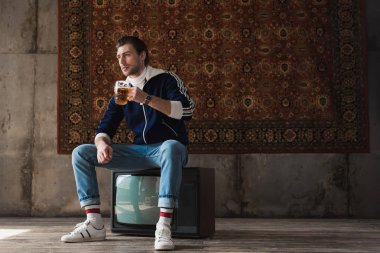 retro üzerinde oturup bira kupa ile vintage giyim düşünceli genç adam tv önünde duvara asılı halı ayarla