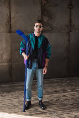 stylish young man in vintage windcheater and sunglasses with hockey stick looking at camera clipart