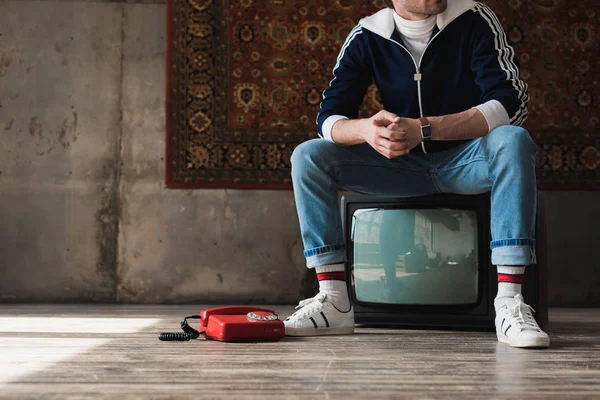 handsome young man in vintage clothes sitting on retro tv set near red wired telephone in front of rug hanging on wall