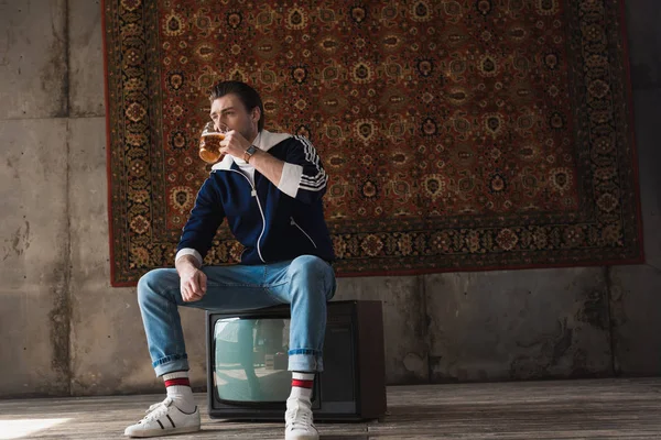 handsome young man in vintage clothes drinking beer while sitting on retro tv set in front of rug hanging on wall