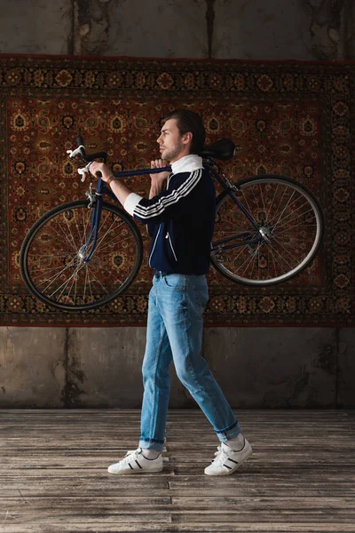 Красивый Молодой Человек Винтажной Одежде Дорожным Велосипедом Плече Перед Ковром — Бесплатное стоковое фото
