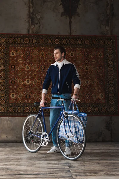 handsome young man in vintage clothes with road bike and bag in front of rug hanging on wall