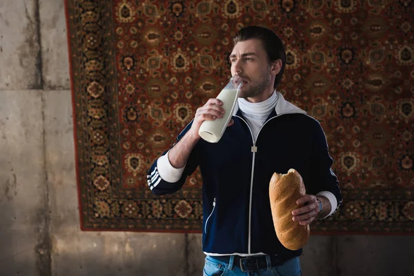 瓶の牛乳と壁に掛かっている敷物の前でパンを一斤とビンテージ服の若い男 — ストック写真