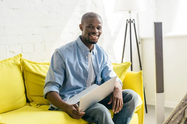 微笑的非洲裔美国人的肖像与笔记本电脑坐在沙发上在家 — 免费的图库照片
