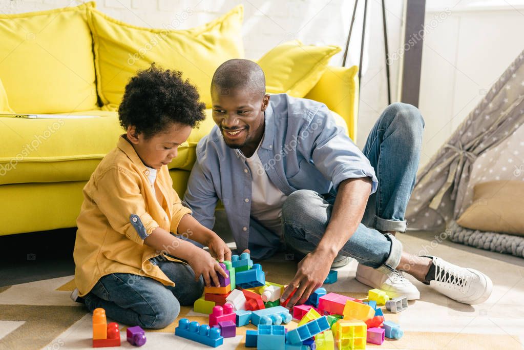 smiling african american father and little son playing with colorful blocks together at home