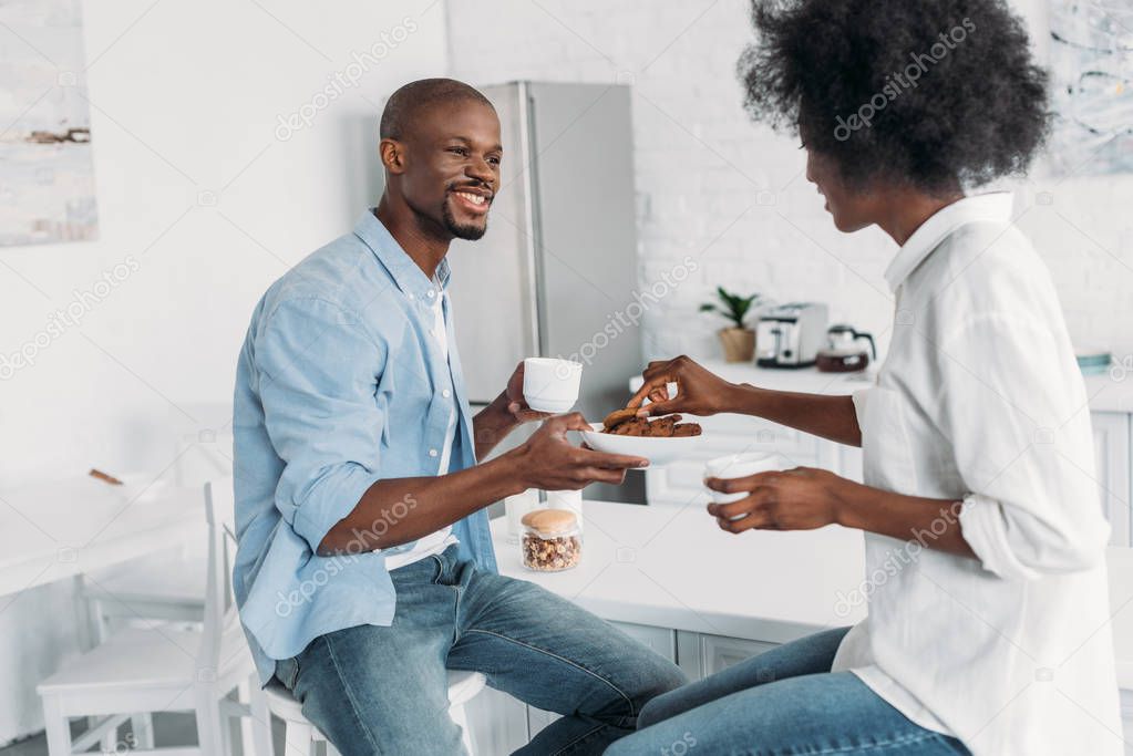african american couple with cups of coffee and cookies in morning together in kitchen at home
