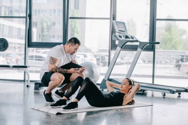 male personal trainer with timer and athletic woman doing abs on fitness mat at gym