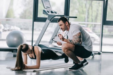 erkek kişisel antrenör gösteren Zamanlayıcı genç atletik kadının tahta spor salonunda fitness mat yapması için