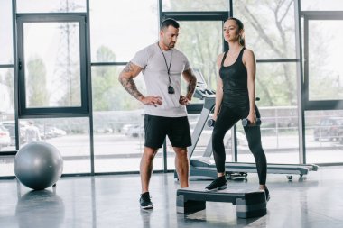 male personal trainer and young sportswoman doing step aerobics exercise with dumbbells at gym clipart