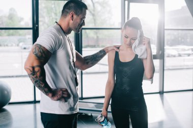 male personal trainer cheering up young sportswoman with bottle of water wiping face with towel at gym clipart