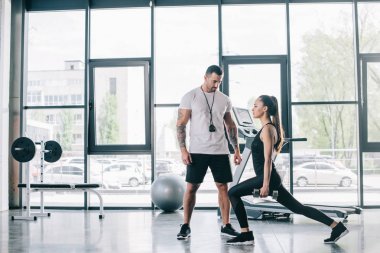 male personal trainer looking at sportswoman doing sit ups with dumbbells at gym clipart