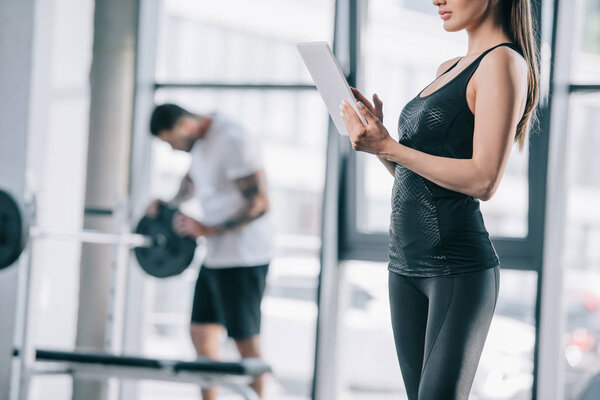 cropped image of female personal trainer with digital tablet and athletic man with barbell at gym