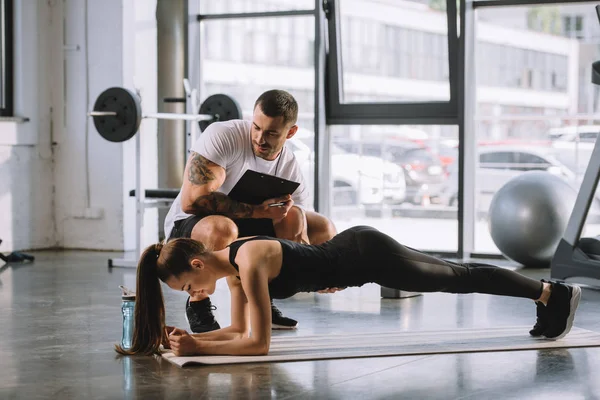 男性私人教练与剪贴板和年轻的运动妇女做木板在健身垫在健身房 — 图库照片