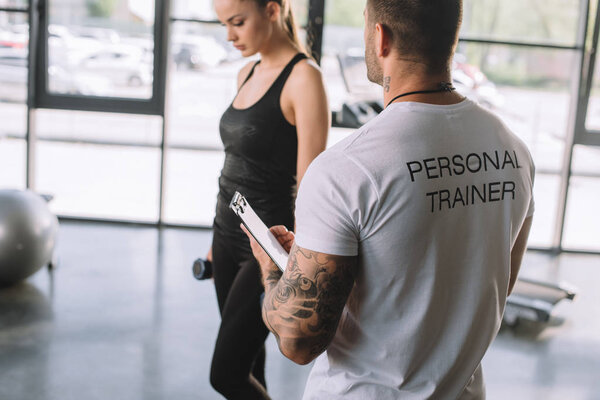 rear view of male personal trainer with clipboard and young sportswoman with dumbbells at gym