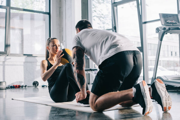 male personal trainer helping sportswoman to do abs with ball at gym