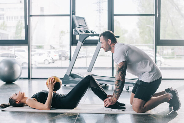 male personal trainer helping sportswoman to do abs with ball at gym