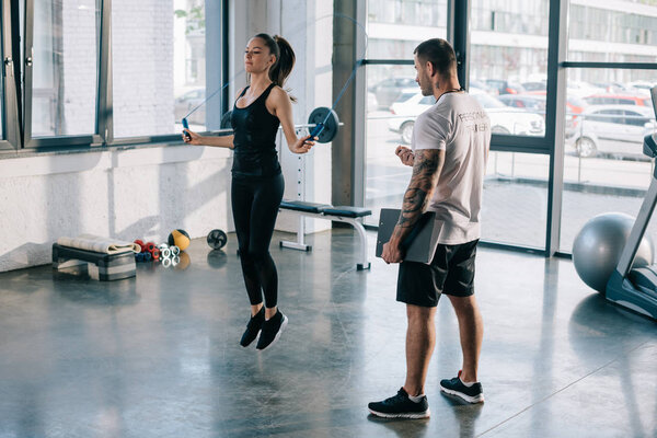 мужчина личный тренер глядя на спортсменку упражнения с скакалкой в тренажерном зале
