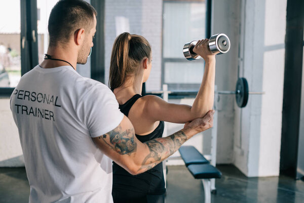 male personal trainer helping sportswoman to do exercises with dumbbell at gym