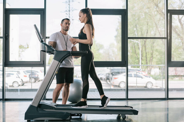 male personal trainer looking at sportswoman on treadmill at gym