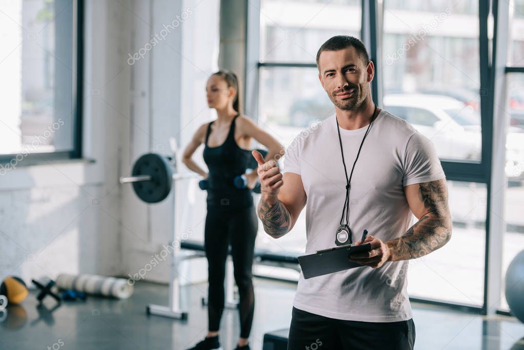 personal trainer with clipboard doing thumb up gesture and young sportswoman exercising with dumbbells behind at gym