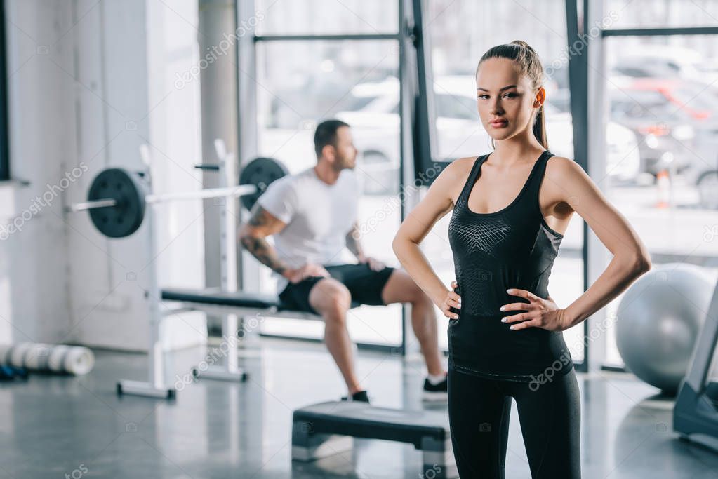 young sportswoman with hands on thighs and man sitting behind at gym