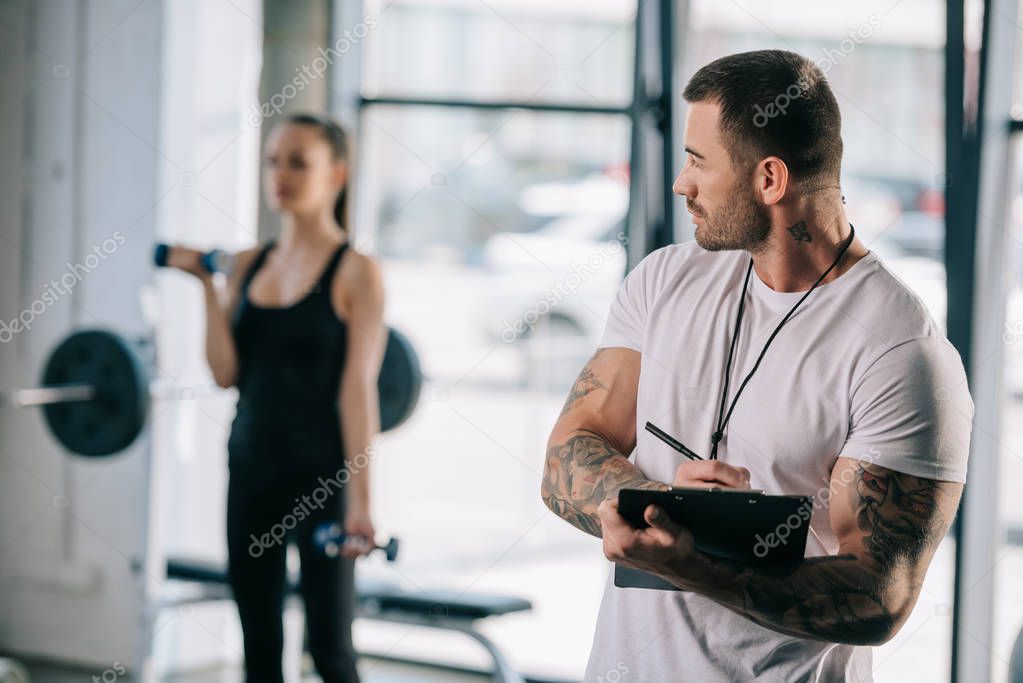 male personal trainer writing in clipboard and young athletic woman exercising with dumbbells behind at gym