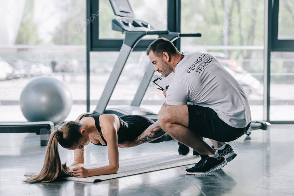 male personal trainer helping young athletic woman to doing plank on fitness mat at gym