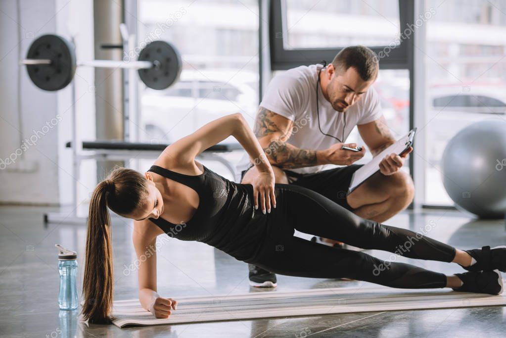 Male personal trainer looking at timer and young athletic woman doing side plank on fitness mat