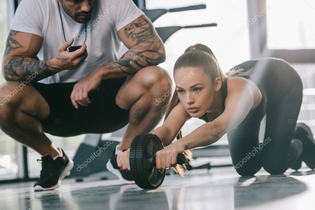 male personal trainer sitting with timer and young sportswoman doing exercise with abs roller at gym