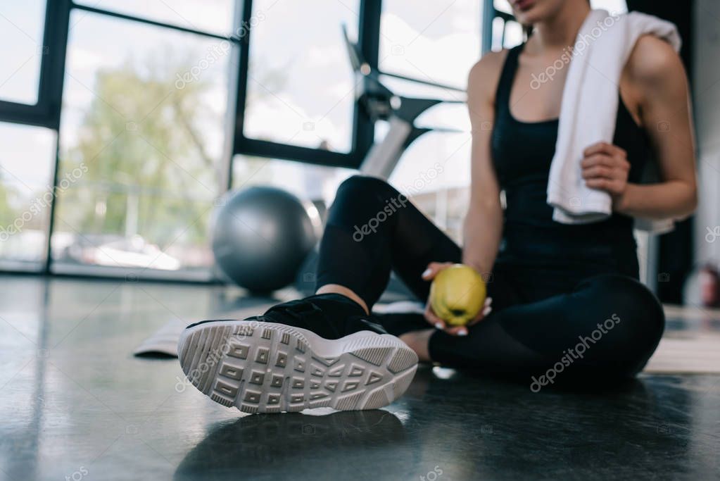 cropped shot of sportswoman resting with towel and apple on gym floor