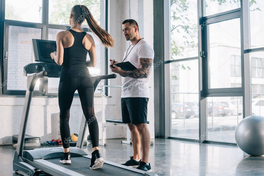 male personal trainer using timer while sportswoman running on treadmill at gym