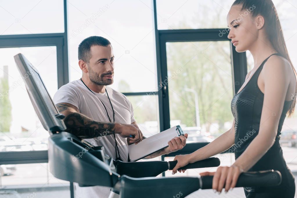 male personal trainer holding clipboard and sportswoman looking at treadmill screen 