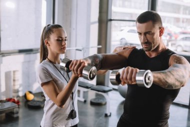 female personal trainer helping sportsman to do exercises with dumbbells at gym clipart