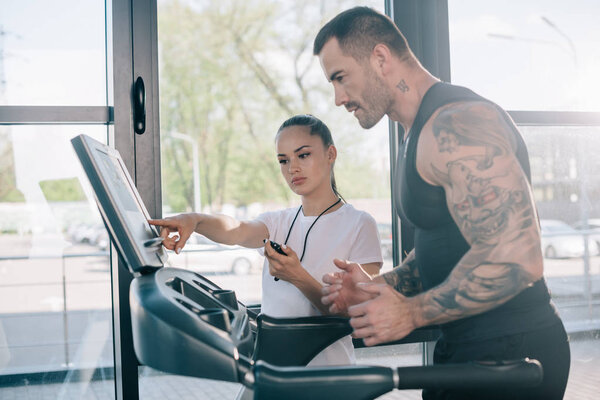 female personal trainer pointing at treadmill screen and running sportsman at gym